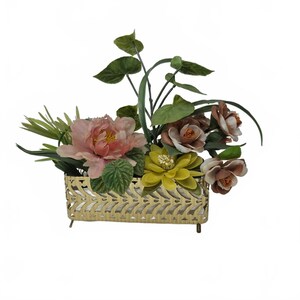 May include: A decorative floral arrangement featuring a variety of artificial flowers in a gold-colored metal container. The arrangement includes pink peonies, yellow and brown flowers, and green foliage. The container has a rectangular shape with a decorative cut-out design.