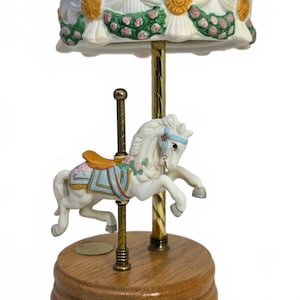 May include: A porcelain carousel music box featuring a white horse with a blue saddle and pink floral garland. The horse is mounted on a gold-toned pole atop a round wooden base. The canopy is white with gold accents and pink floral swags.