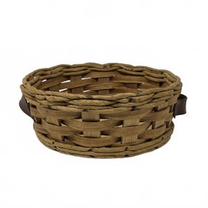 May include: A round, woven basket with a natural brown color. The basket has a sturdy construction with a woven pattern and is accented with dark brown leather handles on each side. The basket is suitable for storage or display.