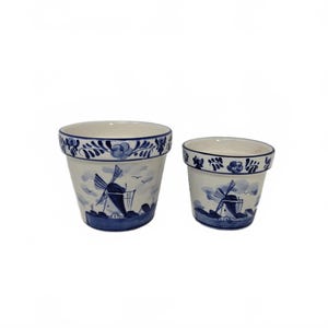 May include: Two ceramic flower pots featuring a Dutch windmill design. The pots are white with blue painted details, including windmills, clouds, and floral patterns around the rims. The pots are different sizes.