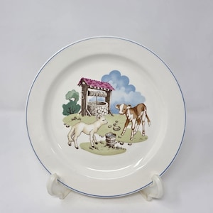May include: A white ceramic plate with a blue rim features a pastoral scene. A lamb and a calf stand near a well with a red roof. The background includes green grass, a blue sky, and a small bucket. The plate is displayed on a white stand.