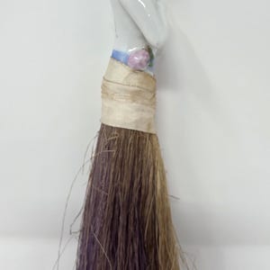 May include: A decorative porcelain figurine of a woman with grey hair and a white dress. The figurine is attached to a brush made of brown and purple fibres, wrapped with beige fabric.