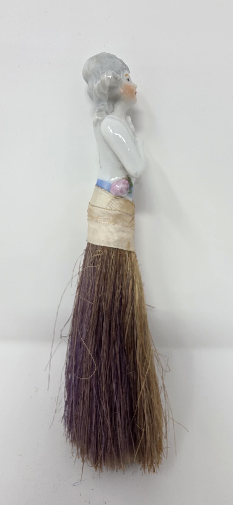 May include: A decorative porcelain figurine of a woman with grey hair and a white dress. The figurine is attached to a brush made of brown and purple fibres, wrapped with beige fabric.