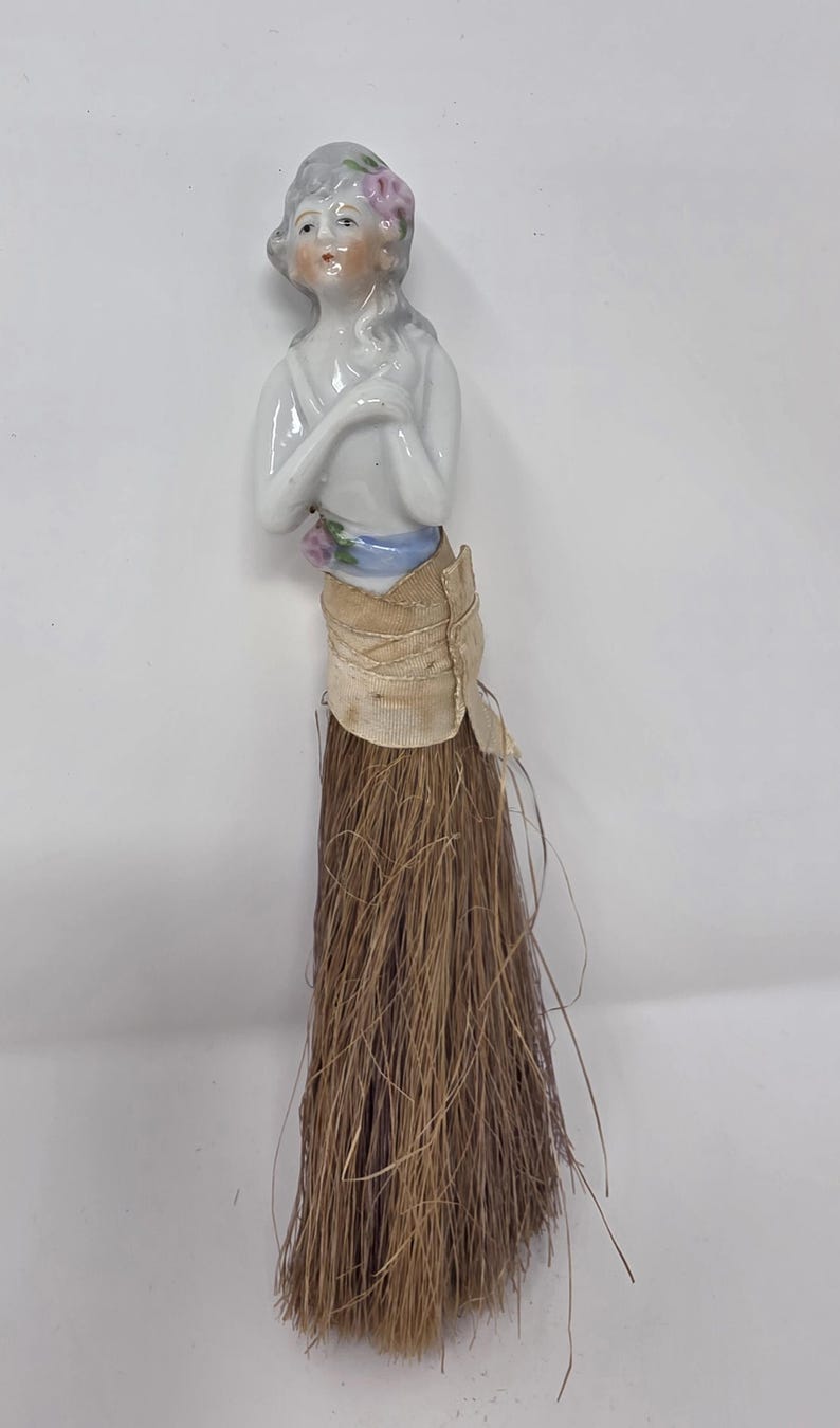 May include: A vintage porcelain doll with a painted face, grey hair, and a pink flower. The doll's torso is attached to a tan fabric skirt and a long, brown, straw-like brush. The doll's arms are crossed.