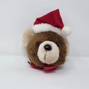May include: A plush teddy bear head with brown fur, tan ears, and a white muzzle. It wears a red Santa hat with white trim and a small red bow. The bear has black eyes.