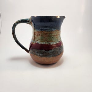 May include: A handcrafted ceramic jug with a dark blue rim, a green and brown mid-section, and a deep red band. The jug has a brown base and a curved handle. The jug is a unique piece of pottery.