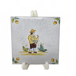 May include: Square ceramic tile with a hand-painted scene of a person playing a drum, wearing a yellow jacket and hat, and brown pants. The tile has a cracked, aged appearance with yellow and green floral accents.