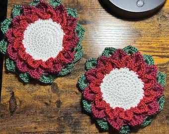 Peut inclure: Deux dessous de verre faits à la main en crochet, en forme de fleur. Chaque dessous de verre présente un centre blanc, entouré de couches de pétales rouges et bordeaux, et bordé de feuilles vertes. Les dessous de verre sont posés sur une surface en bois.