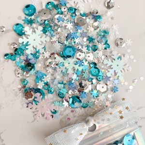 May include: A clear plastic bag filled with a mix of blue, silver, and white snowflake and star-shaped sequins. The bag is tied with a white ribbon with gold stars.
