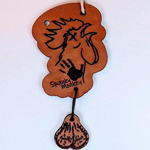 Può includere: Un portachiavi in pelle marrone con un'illustrazione di una scimmia dei cartoni animati e la scritta "Spanky Monkey". Il portachiavi ha un piccolo foro nella parte superiore per una corda e un ciondolo più piccolo attaccato con la scritta "Spanky Monkey".