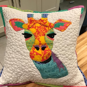 Könnte beinhalten: Ein weißes Kissen mit einer bunten Giraffenkopf-Applikation. Das Giraffenkopf ist aus verschiedenen gemusterten Stoffen in Grün-, Gelb-, Orange-, Rosa- und Violetttönen gefertigt. Das Kissen hat einen weißen Hintergrund mit einem gesteppten Muster.
