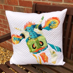 Pode incluir: Uma almofada branca com um design de vaca de patchwork colorido. O rosto da vaca é costurado com tecido azul brilhante, verde, amarelo e laranja. A almofada está sentada em um banco de madeira.