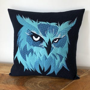May include: A blue and black patterned owl cushion cover. The owl's eyes are white with black pupils. The cushion cover is made of fabric and has a quilted texture.