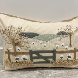 Può includere: Un cuscino beige con una scena ricamata di una campagna innevata con un cancello, alberi e pecore. Il ricamo è realizzato in tonalità di marrone, verde e bianco.