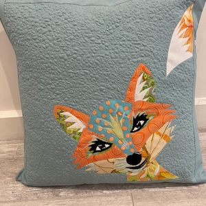 May include: A blue pillow with a fox face made of colorful fabric scraps. The fox's eyes are black and white, and its nose is black. The fox's fur is orange, green, and blue.