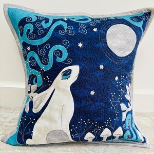 May include: A blue and white decorative pillow with a white rabbit looking up at a full moon. The pillow features a whimsical design with stars, swirls, and mushrooms.