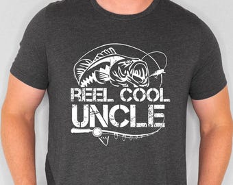 Puede incluir: Una camiseta gris oscuro con un gráfico blanco de un pez y el texto "Reel Cool Uncle".