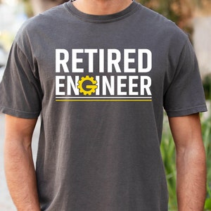 May include: A dark gray t-shirt with the text "RETIRED ENGINEER" in white letters. A yellow gear symbol is incorporated into the word "ENGINEER".