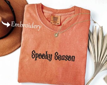 May include: An orange-red t-shirt with the embroidered text "Spooky Season" in black. A gold necklace with a pendant is draped across the shirt. A brown hat is visible in the background.