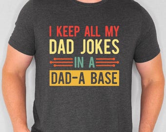 May include: Dark gray t-shirt with the text "I KEEP ALL MY DAD JOKES IN A DAD-A BASE" in red, yellow, and teal. The shirt is a crew neck style and has short sleeves.