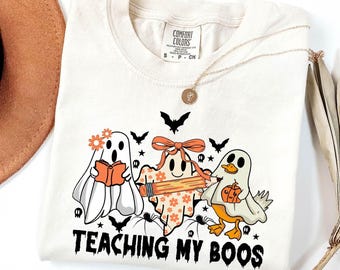May include: Cream-colored t-shirt featuring a Halloween-themed graphic. The design includes three cartoon ghosts, bats, spiders, and the text "TEACHING MY BOOS". One ghost reads a book, another holds a pencil, and the third holds a pumpkin.