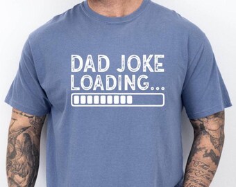 May include: A blue t-shirt with the text "DAD JOKE LOADING..." in white, along with a loading bar graphic. The shirt is a solid color and appears to be made of a soft material.