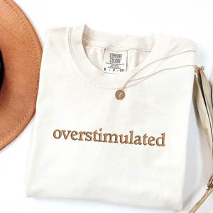 May include: A cream coloured t-shirt with the words "overstimulated" embroidered in brown on the front. The shirt is lying flat on a white surface.