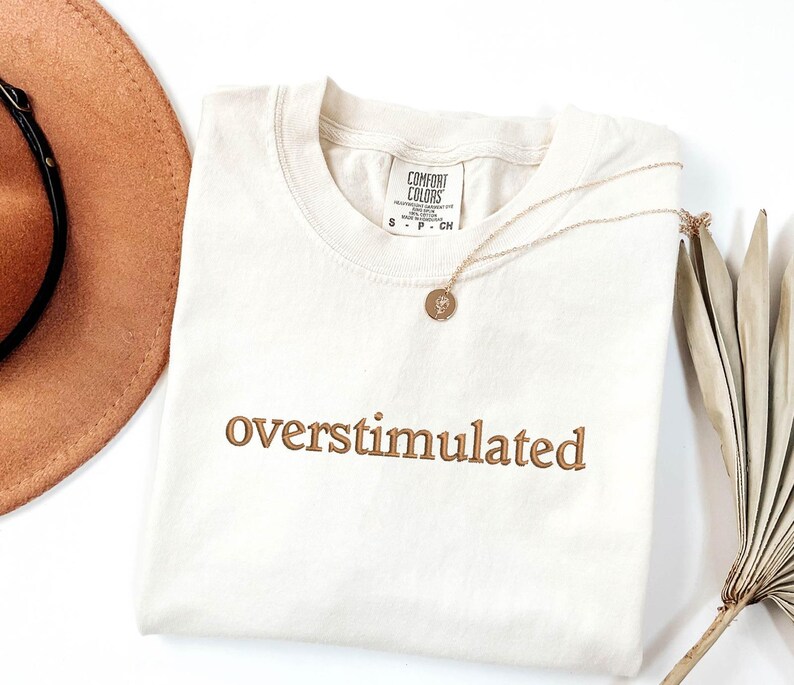 May include: A cream coloured t-shirt with the words "overstimulated" embroidered in brown on the front. The shirt is lying flat on a white surface.