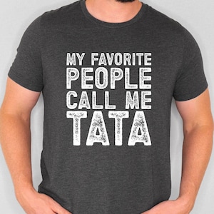 May include: Dark gray t-shirt with the white text "MY FAVORITE PEOPLE CALL ME TATA". The text is in a bold, blocky font. The shirt has short sleeves and a crew neck.