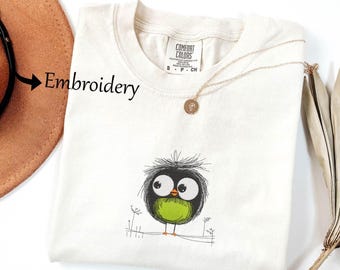 May include: Cream-colored Comfort Colors t-shirt with an embroidered cartoon bird design. The bird has large eyes, a green belly, and a black and gray outline. A gold necklace with a pendant is draped over the shirt. A brown hat is visible in the background.
