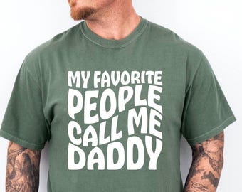 May include: A green t-shirt with the text "MY FAVORITE PEOPLE CALL ME DADDY" in white, bold, bubble letters. The shirt has short sleeves and a crew neck. The shirt is made of a soft, comfortable material.