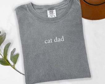 Può includere: Maglietta grigia con il testo "cat dad" ricamato in bianco. La maglietta è del marchio Comfort Colors ed è di taglia L.