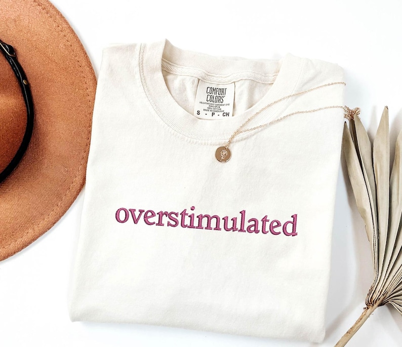 May include: A cream coloured t-shirt with the word "overstimulated" embroidered in pink on the front. The shirt is lying flat on a white surface.