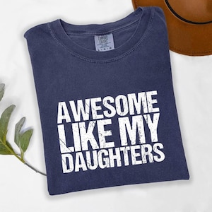 May include: A navy blue short-sleeved t-shirt with a distressed white graphic print on the front that reads "AWESOME LIKE MY DAUGHTERS" in capital letters. The shirt features a crew neckline and a Comfort Colors brand label at the inner collar.