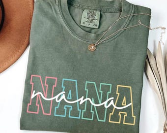 May include: An olive green t-shirt with the word "NANA" in a colorful, block letter design. The word "nana" is written in a cursive font over the block letters. A gold necklace with a pendant is draped over the shirt.