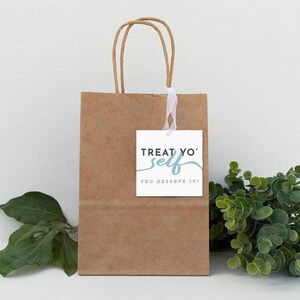 Op de afbeelding: Een bruine papieren cadeauzak met een wit cadeau-etiket waarop staat "Treat Yo' Self You Deserve It!" De tas is vastgebonden met een wit lint en er is een kleine groene plant op de achtergrond.