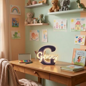 Può includere: Una lampada a forma di lettera G blu scuro, illuminata con il nome Genny in corsivo bianco caldo. La lampada è posizionata su una scrivania in legno accanto a una scatola di metallo con matite colorate, due libri e una piccola pianta succulenta.