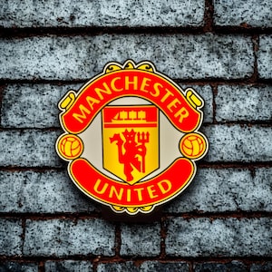 May include: A circular Manchester United logo, featuring a red and yellow color scheme. The logo has the words "Manchester United" in a red banner, with a yellow shield in the center. The shield has a red devil and a ship.
