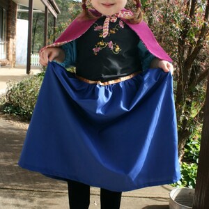 Puede incluir: Un disfraz infantil con una falda azul, un corpi&ntilde;o negro con bordado floral y una capa morada. El disfraz incluye un cintur&oacute;n dorado y se combina con botas negras. El atuendo est&aacute; dise&ntilde;ado para disfrazarse y jugar con imaginaci&oacute;n.
