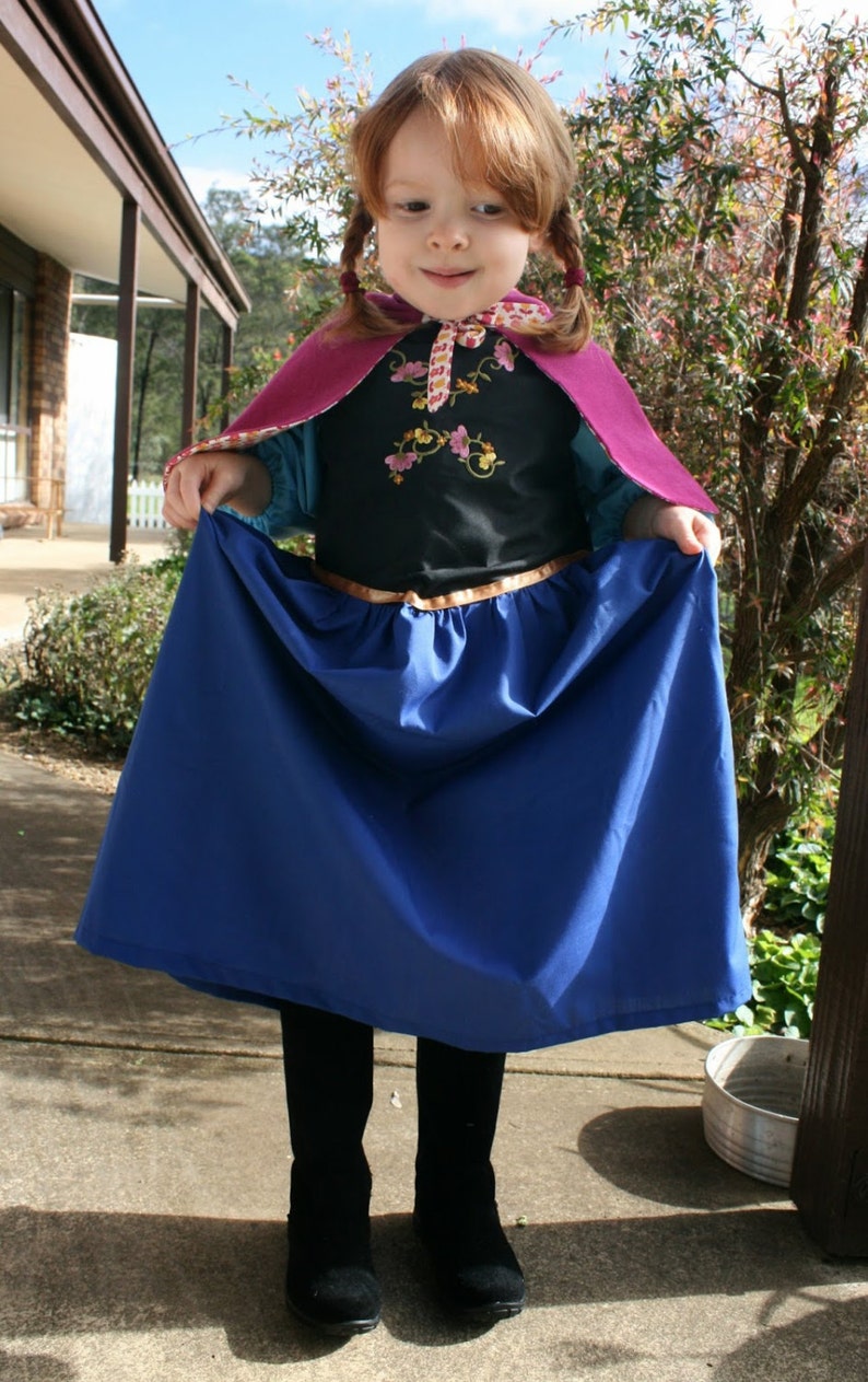Puede incluir: Un disfraz infantil con una falda azul, un corpi&ntilde;o negro con bordado floral y una capa morada. El disfraz incluye un cintur&oacute;n dorado y se combina con botas negras. El atuendo est&aacute; dise&ntilde;ado para disfrazarse y jugar con imaginaci&oacute;n.