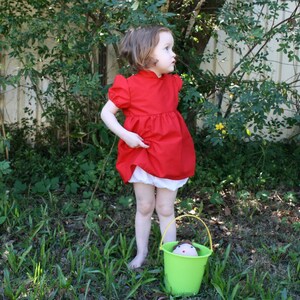 Puede incluir: Una ni&ntilde;a peque&ntilde;a con un vestido rojo brillante de manga corta y falda fruncida, combinado con bragas blancas. La ni&ntilde;a est&aacute; descalza en una zona de c&eacute;sped, sujetando el vestido. Un cubo verde con una mu&ntilde;eca est&aacute; cerca.
