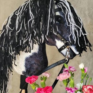 May include: Black and white pinto horse hobby horse with a black leather bridle and reins. The horse has a long black and white mane and tail. The horse is standing on a wooden stick with a brown leather strap.