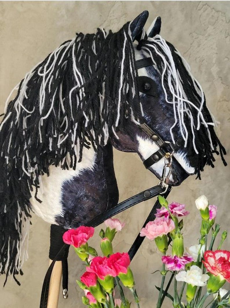 May include: Black and white pinto horse hobby horse with a black leather bridle and reins. The horse has a long black and white mane and tail. The horse is standing on a wooden stick with a brown leather strap.