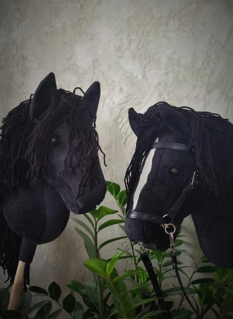 May include: Two black hobby horse heads with black yarn manes. One horse has a white stripe on its face and a black leather bridle.