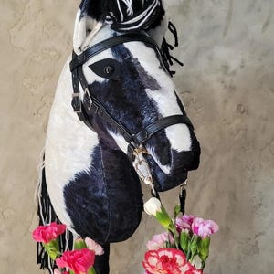 May include: Black and white horse head with a black leather bridle and a black and white mane. The horse head is surrounded by pink and white carnations.