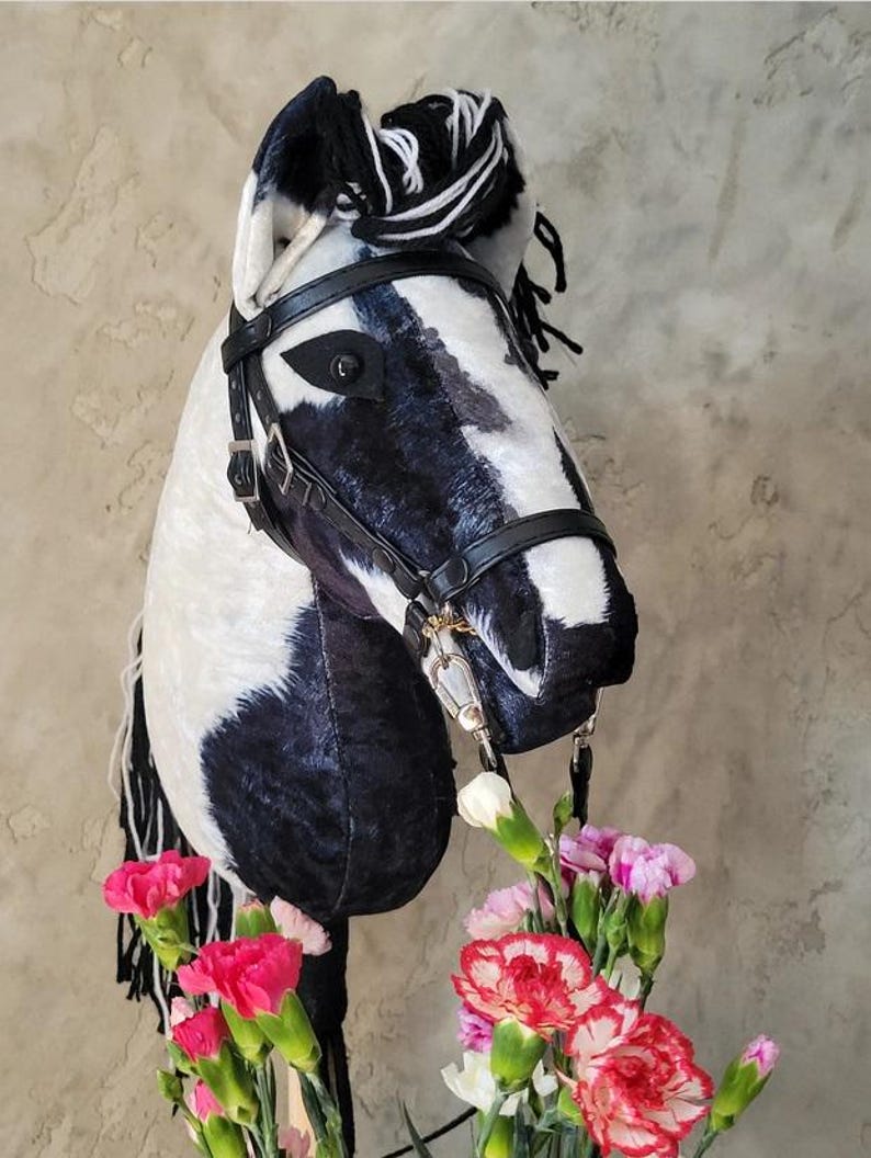 May include: Black and white horse head with a black leather bridle and a black and white mane. The horse head is surrounded by pink and white carnations.