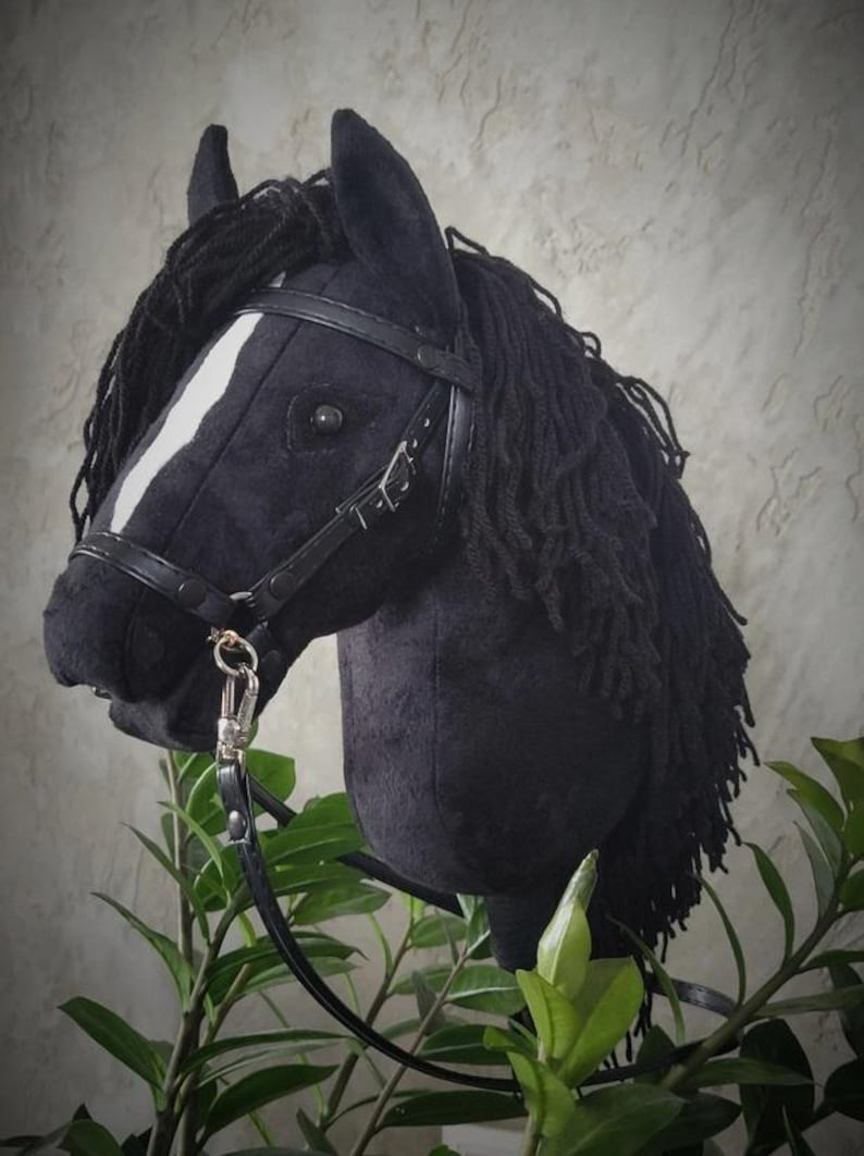 May include: Black horse head with a white stripe on its face, wearing a black leather bridle with a silver buckle. The horse head is made of plush fabric and has a long black mane.