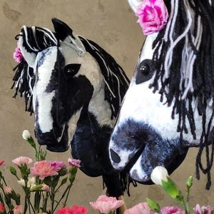 May include: Two black and white horse head rocking horse toys with pink flowers on their manes. The horses are made of fabric and have black yarn for manes and tails.