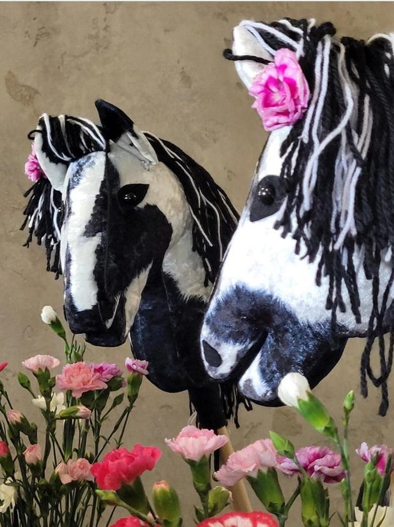 May include: Two black and white horse head rocking horse toys with pink flowers on their manes. The horses are made of fabric and have black yarn for manes and tails.