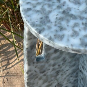 May include: A white and gray speckled round side table with a cutout design on the side. The table is made of recycled plastic and is perfect for outdoor use.